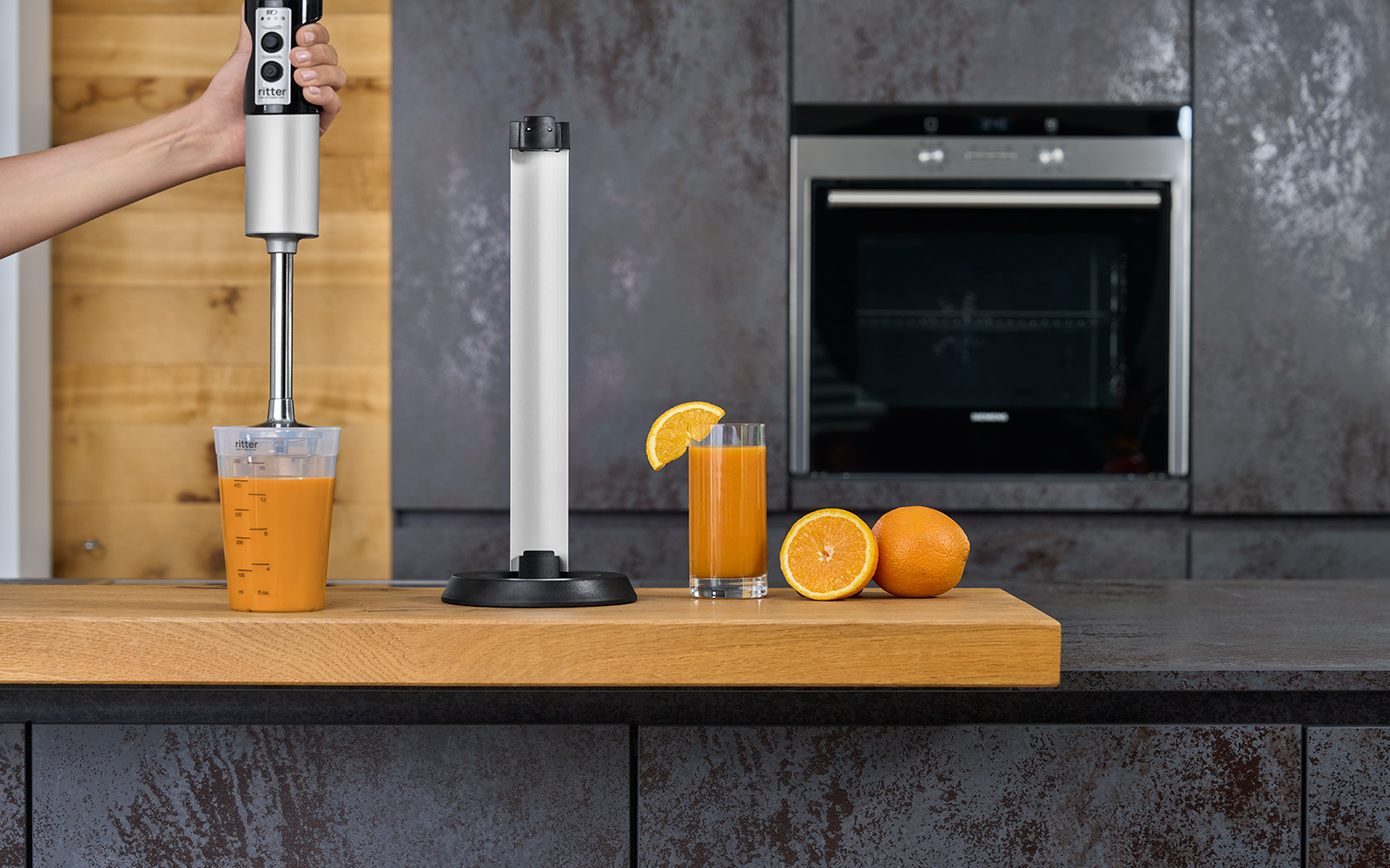A picture of a silver hand blender blending oranges in a measuring jug and standing in a kitchen next to its charging station.