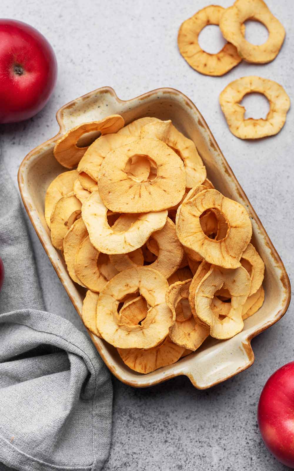 Schale mit knusprigen, goldbraunen Apfelchips in runder Form, daneben frische rote Äpfel und ein graues Tuch Schale mit knusprigen, goldbraunen Apfelchips in runder Form, daneben frische rote Äpfel und ein graues Tuch