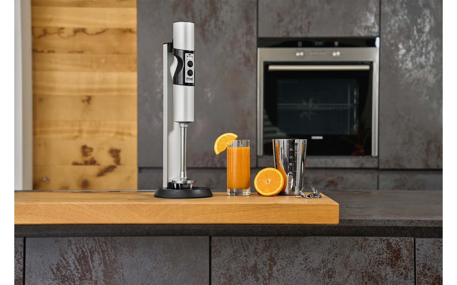A picture of a silver hand blender standing next to a glass of orange juice in a kitchen.