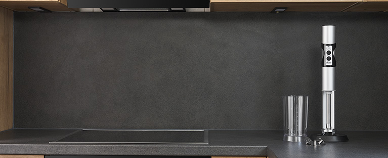 Ein Bild von einem silbernen Stabmixer, einem gefüllten Messbecher und einem, mit Saft gefüllten, Glas, in einer Küche.