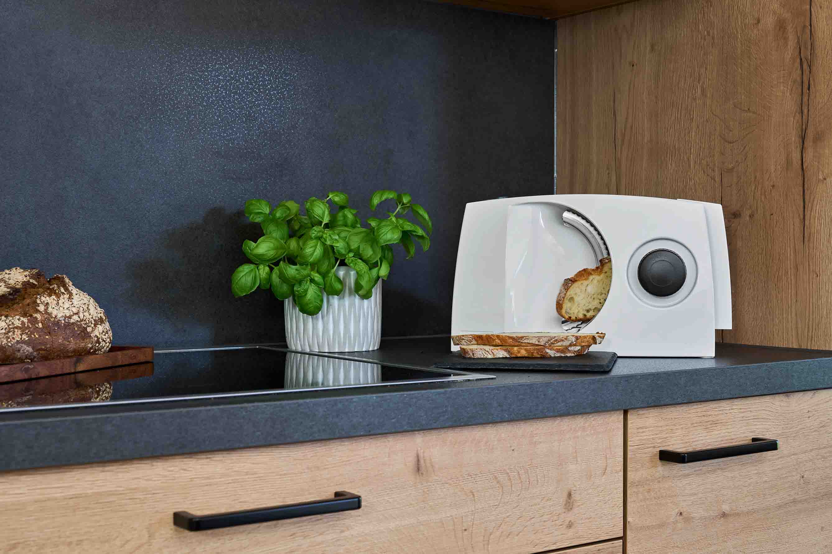 Weißer Allesschneider auf dunkler Küchenarbeitsplatte mit geschnittenem Schneidgut und einem Topf Basilikum.