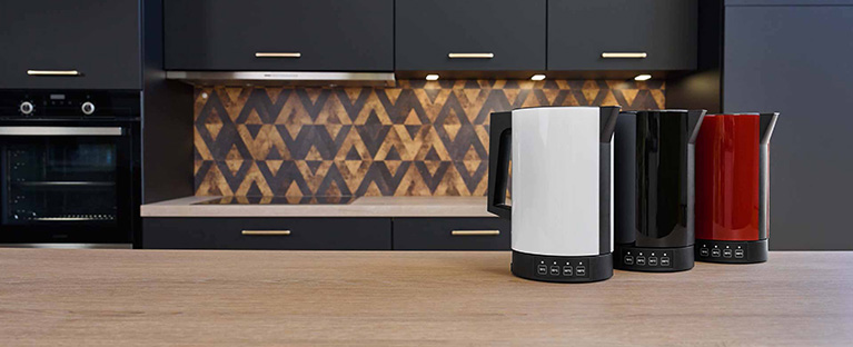 Ein Bild von einem roten, einem schwarzen und einem weißen Wasserkocher die nebeneinander auf einer Küchenplattform stehen.