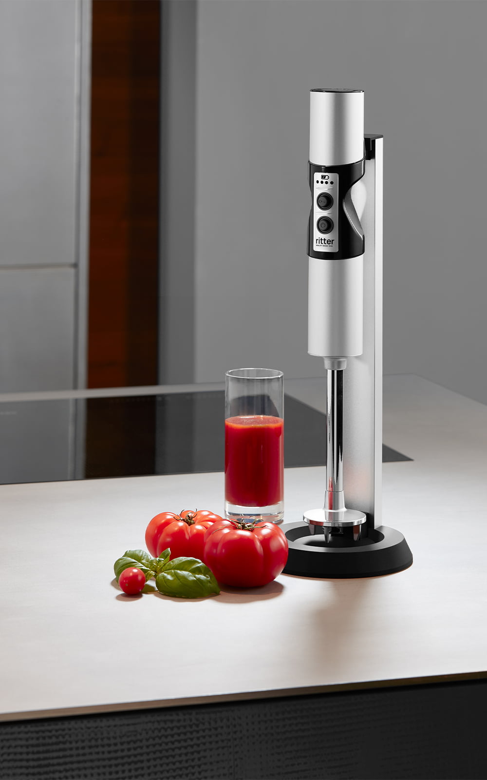 A picture of a silver hand blender standing next to a glass of tomato juice in a kitchen. A picture of a silver hand blender standing next to a glass of tomato juice in a kitchen.