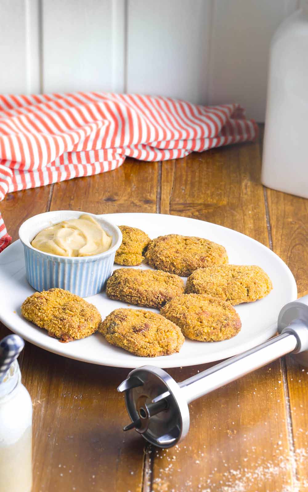 Ein Teller mit knusprigen, goldbraun gebackenen Veggie-Nuggets, serviert mit einer Schale cremigem Dip. Daneben liegt ein Stabmixer, im Hintergrund ein rot-weiß gestreiftes Tuch auf einem Holztisch Ein Teller mit knusprigen, goldbraun gebackenen Veggie-Nuggets, serviert mit einer Schale cremigem Dip. Daneben liegt ein Stabmixer, im Hintergrund ein rot-weiß gestreiftes Tuch auf einem Holztisch