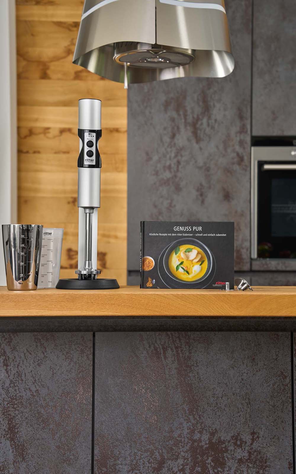A picture of a silver hand blender standing next to a recipe book in a kitchen. A picture of a silver hand blender standing next to a recipe book in a kitchen.