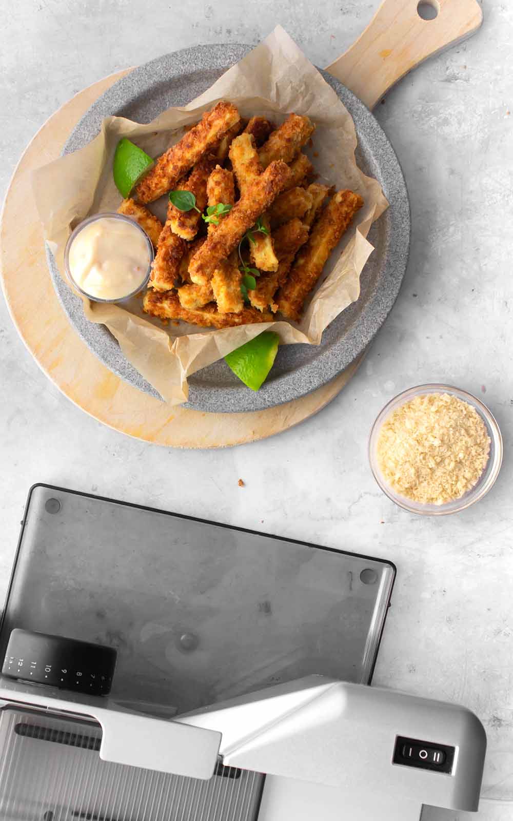Knusprig gebackene Tofu-Sticks auf Backpapier, serviert auf einem Teller mit Dip und Limettenstück. Daneben eine kleine Schale mit Panko-Bröseln und im Vordergrund eine silberne Küchenmaschine Knusprig gebackene Tofu-Sticks auf Backpapier, serviert auf einem Teller mit Dip und Limettenstück. Daneben eine kleine Schale mit Panko-Bröseln und im Vordergrund eine silberne Küchenmaschine