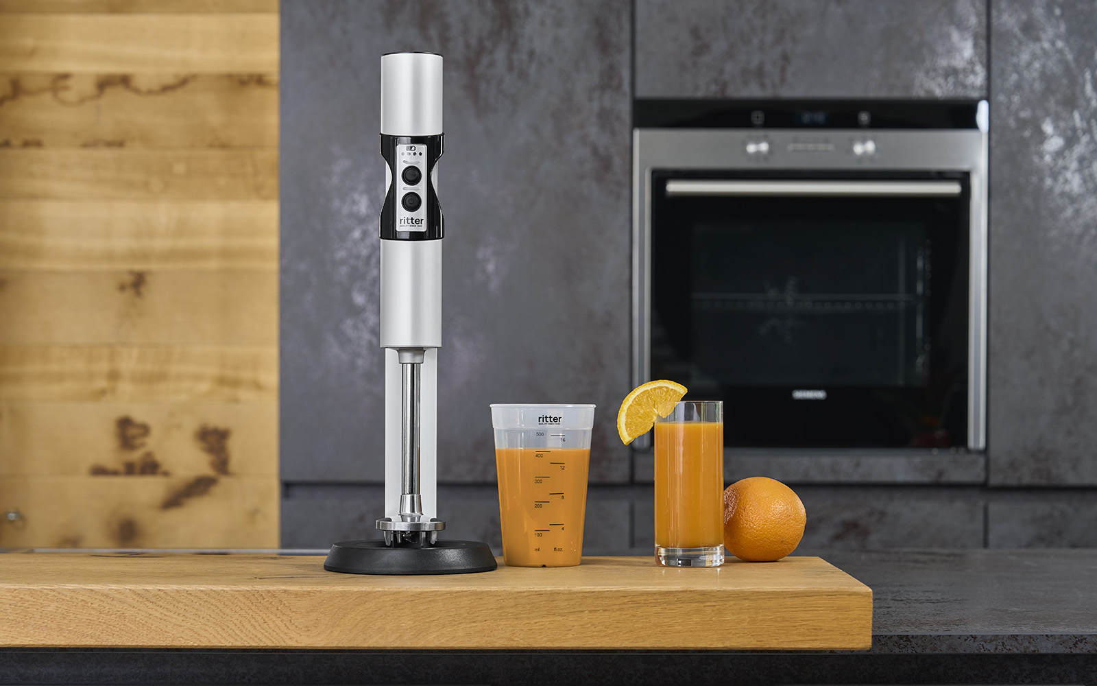 A picture of a silver hand blender, a filled measuring jug and a glass filled with juice in a kitchen.
