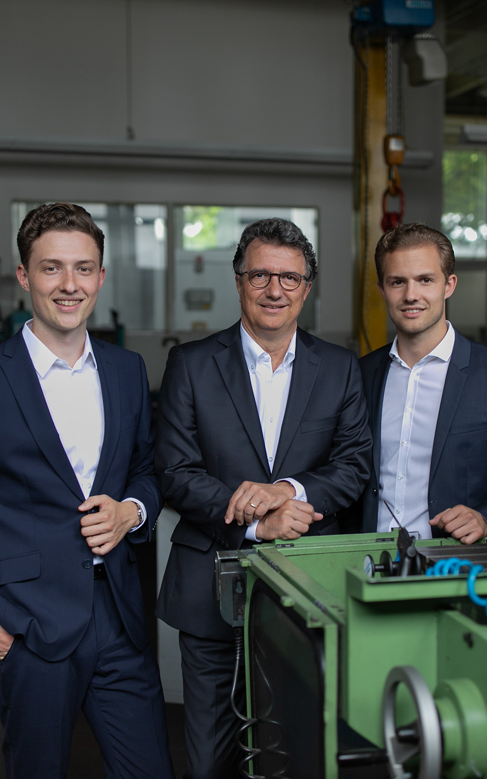 A picture of three men in suits standing in a workshop and smiling at the camera. A picture of three men in suits standing in a workshop and smiling at the camera.