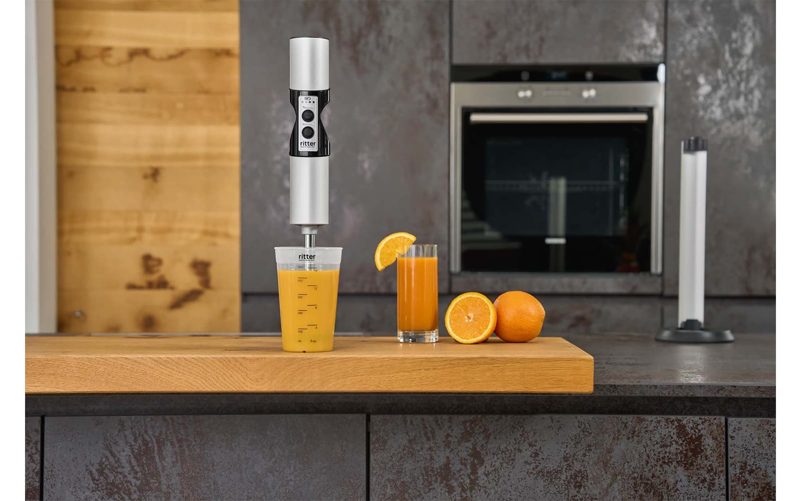 A picture of a silver hand blender blending oranges in a measuring jug and standing in a kitchen.