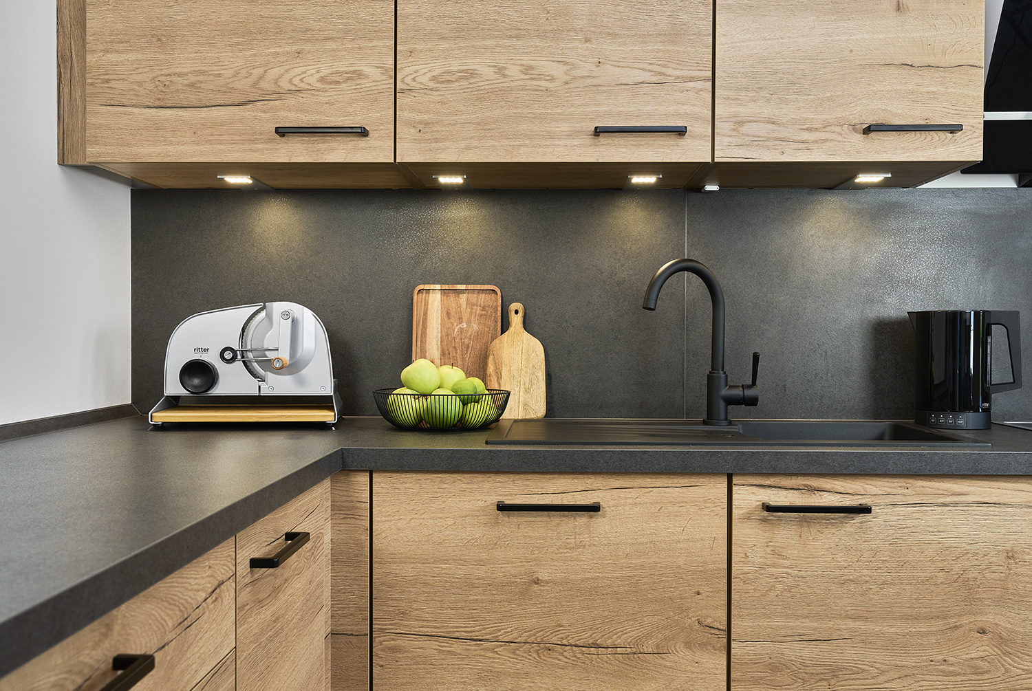 Silberner manueller Allesschneider der Marke ritter mit schwarzem Drehknopf, Griff und Bambusbrett, auf Arbeitsplatte mit einer Schale mit Äpfeln rechts daneben in moderner Küche