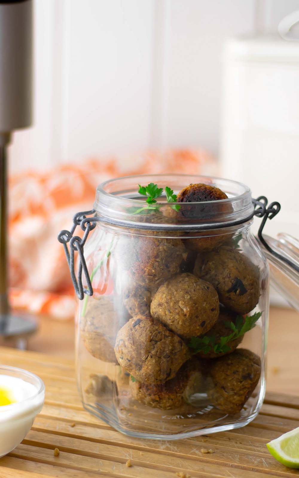 Ein Glas mit selbstgemachten veganen Falafel-Bällchen, garniert mit frischer Petersilie. Im Vordergrund steht eine kleine Schale mit Dip, daneben ein Limettenstück. Im Hintergrund ein Stabmixer und ein orange-weißes Küchentuch auf einer Holzoberfläche Ein Glas mit selbstgemachten veganen Falafel-Bällchen, garniert mit frischer Petersilie. Im Vordergrund steht eine kleine Schale mit Dip, daneben ein Limettenstück. Im Hintergrund ein Stabmixer und ein orange-weißes Küchentuch auf einer Holzoberfläche